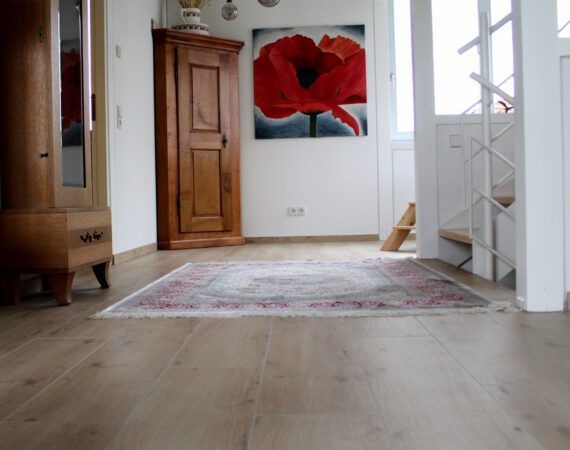 Holzschrank, darüber ein Gemälde mit roter Mohnblume, über einem gemusterten Teppich auf Holzboden.