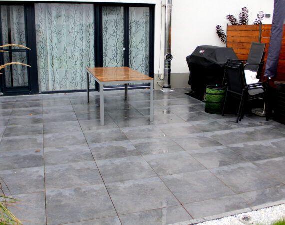 Grau geflieste Terrasse mit Tisch, Grill, Stühlen und Glasschiebetüren.