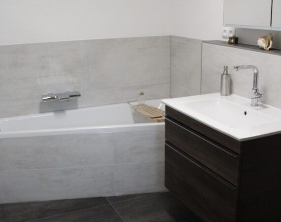 Modernes Badezimmer mit Badewanne, Waschbecken und dunklem Holzwaschtisch. Wände und Boden sind grau gefliest.