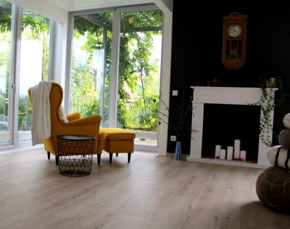 Gemütliches Wohnzimmer mit gelbem Sessel, kleinem Tisch und Hocker. Weiß umrahmter Kamin, große Fenster mit Blick ins Grüne.