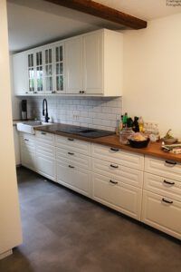 Weiße Küche mit Oberschränken, langer Arbeitsplatte, Backstein-Spritzschutz und dunklen Beschlägen.