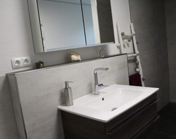 Modernes Badezimmer mit weißem Waschbecken, grauen Schränken, Spiegel und Handtuchhalter.