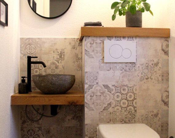 Kleines Badezimmer mit gemusterten Fliesen, schwarzem Wasserhahn, Steinwaschbecken, Holzregalen und Toilette.