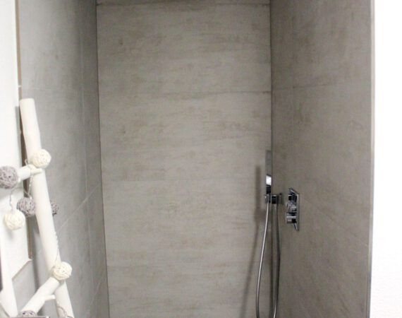 Grau geflieste Dusche mit Duschkopf, Wasserhahn und dekorativem Ablagefach auf der linken Seite.