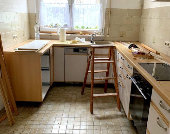 Die Küche wird renoviert. Schränke, Arbeitsplatten, Spüle und Elektrogeräte sind sichtbar. Eine Leiter ist vorhanden.