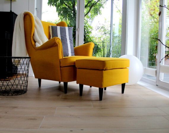 Gelber Sessel mit Hocker am Fenster. Eine weiße Decke und ein gestreiftes Kissen auf dem Sessel. Heller Holzboden.