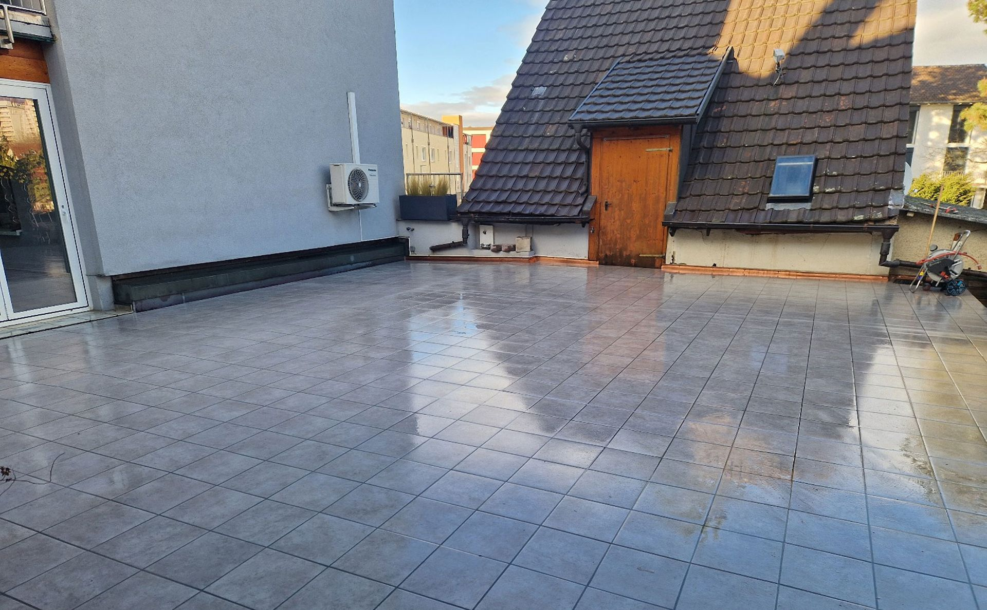 Grau geflieste Dachterrasse mit Holztür und einem modernen Gebäude im Hintergrund.