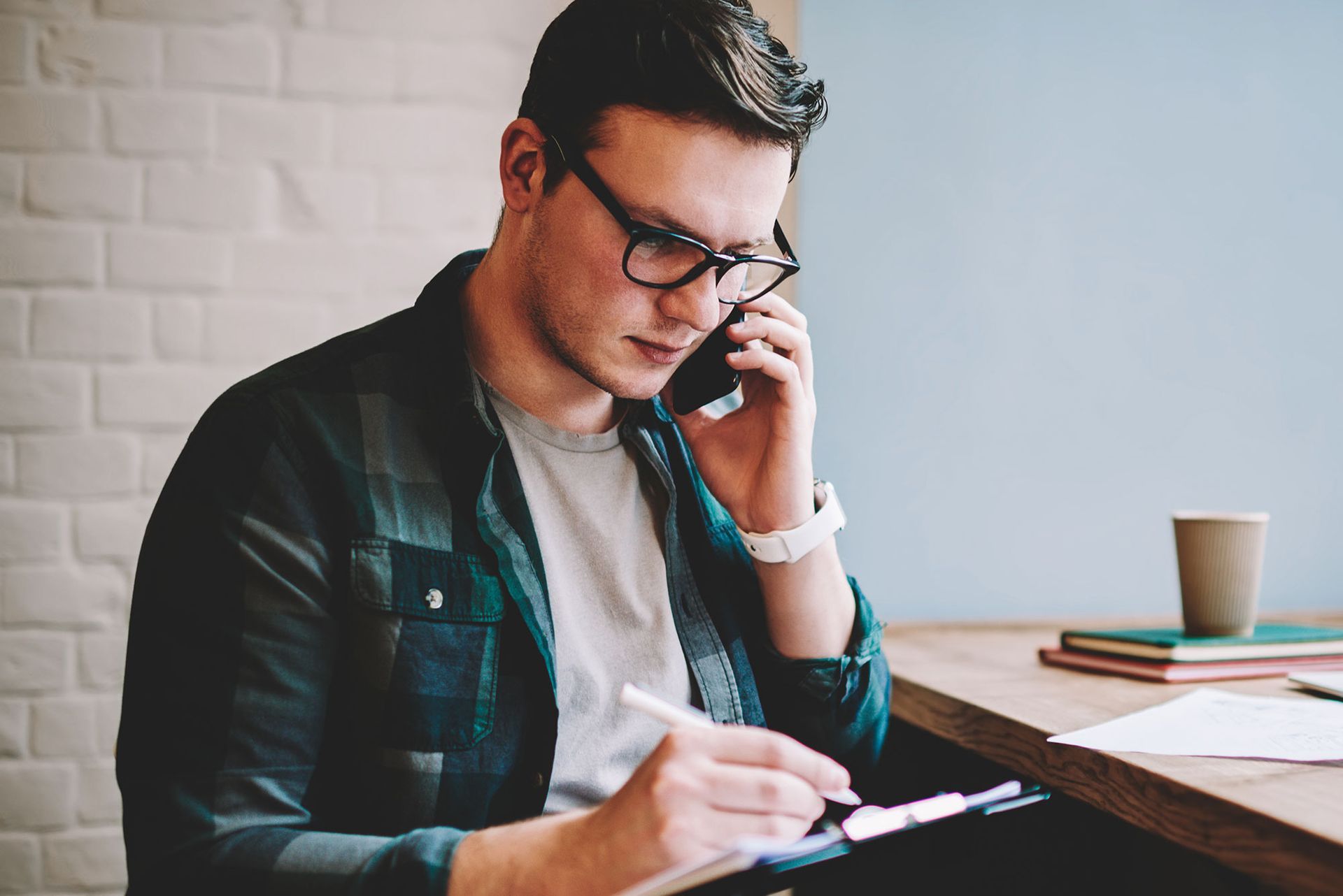 Mann mit Brille und kariertem Hemd, der an einem Tisch telefoniert und sich Notizen macht.