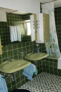 Badezimmer mit grünen Fliesen, zwei Waschbecken, Wandschrank, Spiegel und Duschvorhang.