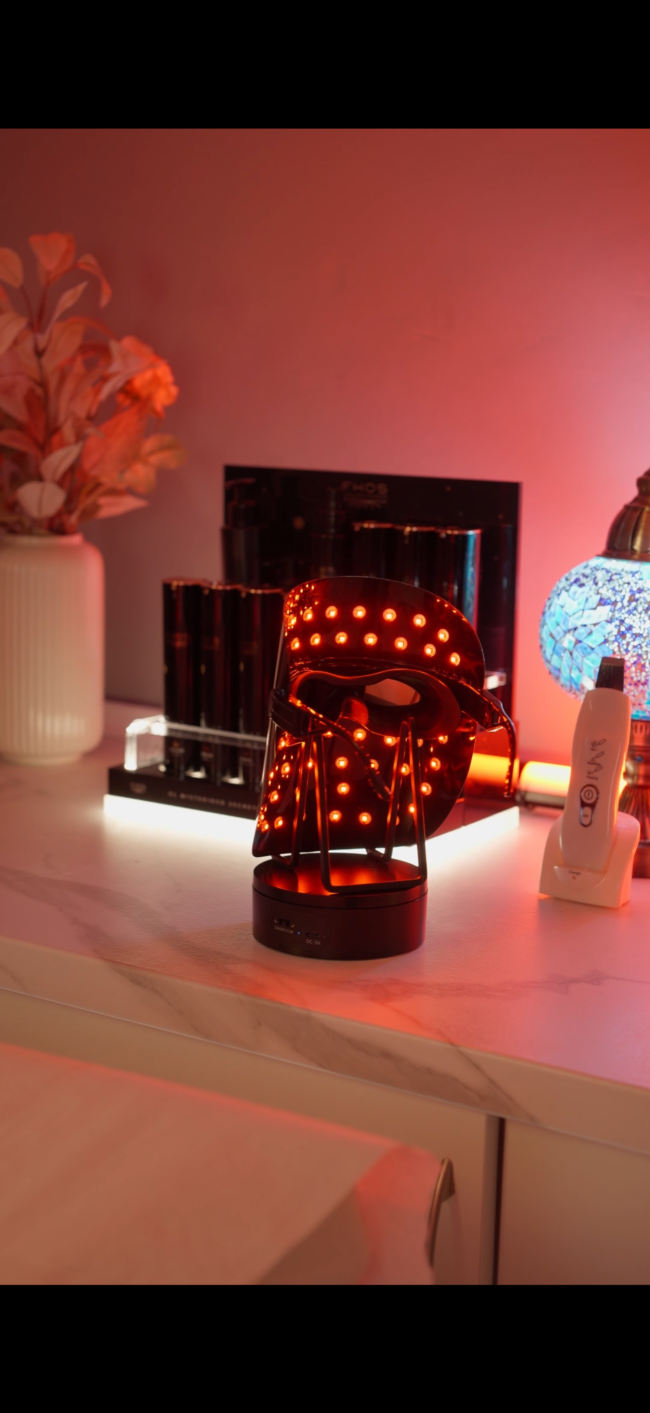 Mesa iluminada en rojo con una lámpara estampada brillante, un jarrón con flores secas y una pieza decorativa azul texturizada.