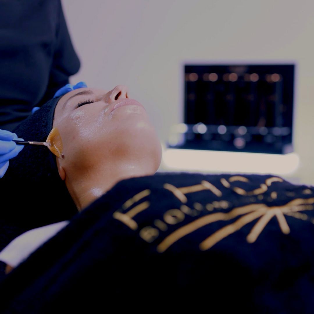 Persona que recibe un tratamiento facial cosmético mientras está recostada, con una mano enguantada sosteniendo un instrumento cerca del rostro.