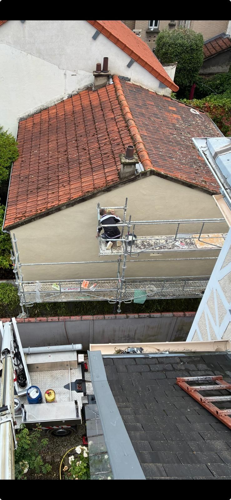 Vue aérienne d'une maison au toit de tuiles rouges, d'ouvriers sur un échafaudage et des toits environnants.