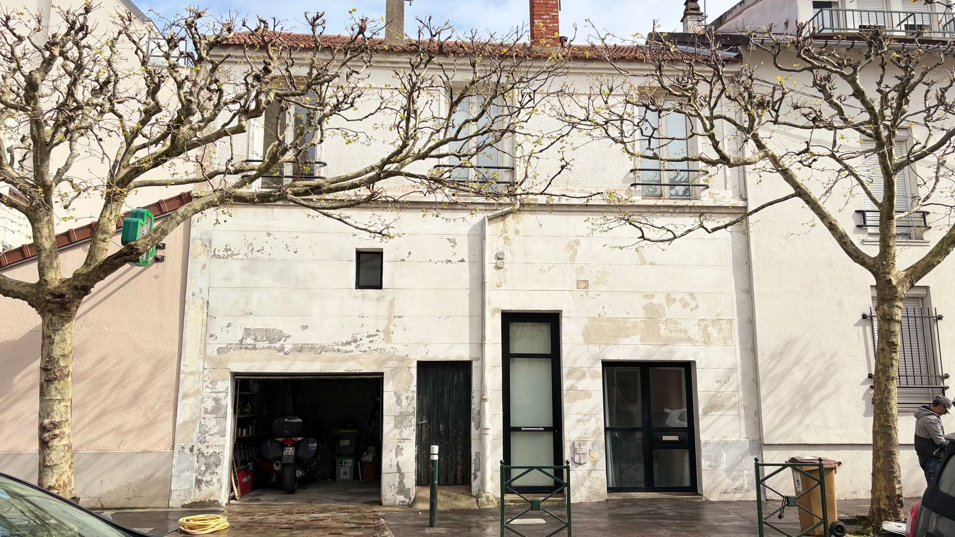Vieux bâtiment blanc avec des arbres dénudés devant et une porte ouverte ressemblant à celle d'un garage.