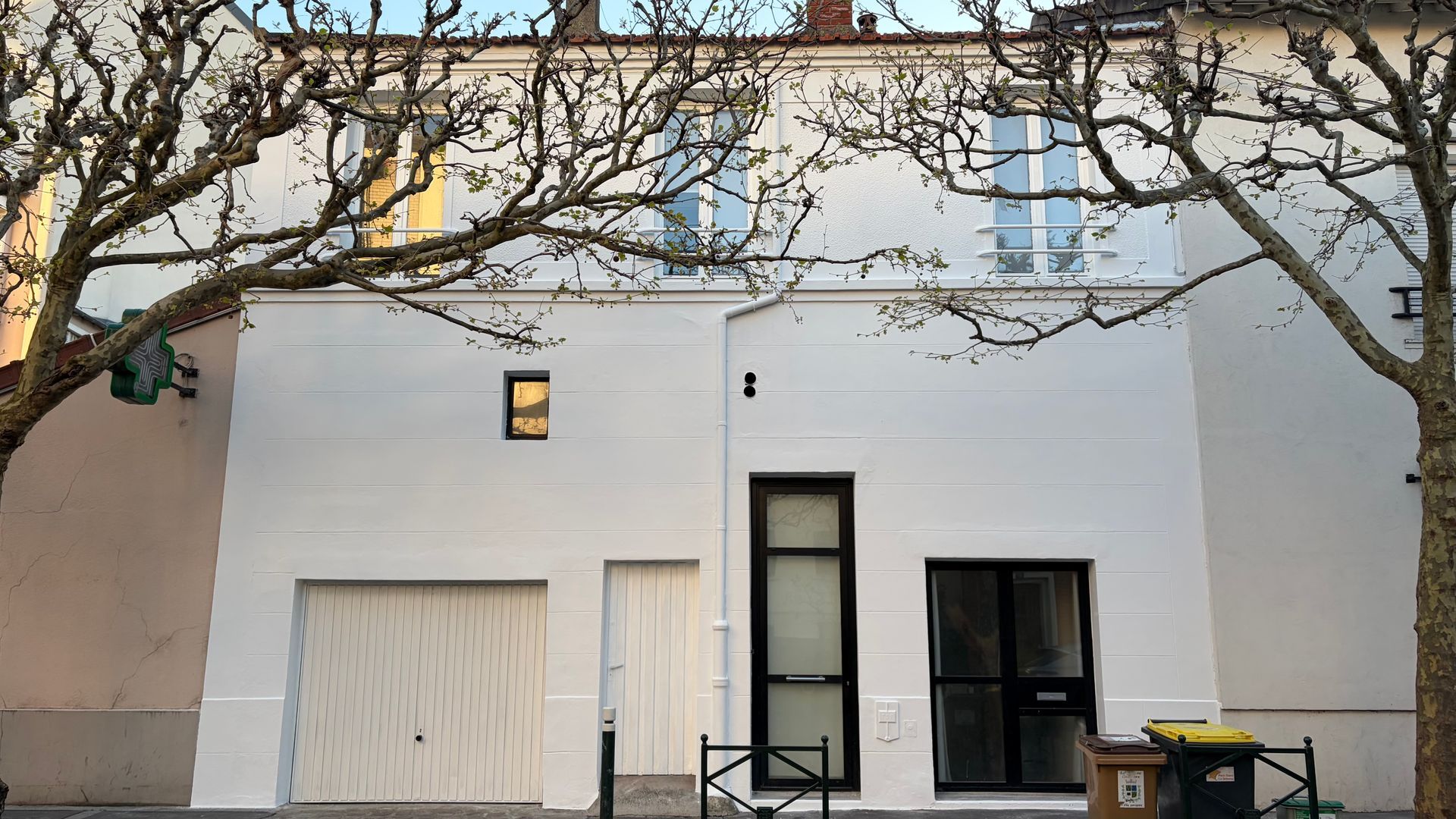 Bâtiment blanc de deux étages avec des branches d'arbres dénudées devant, porte de garage et porte d'entrée noire.
