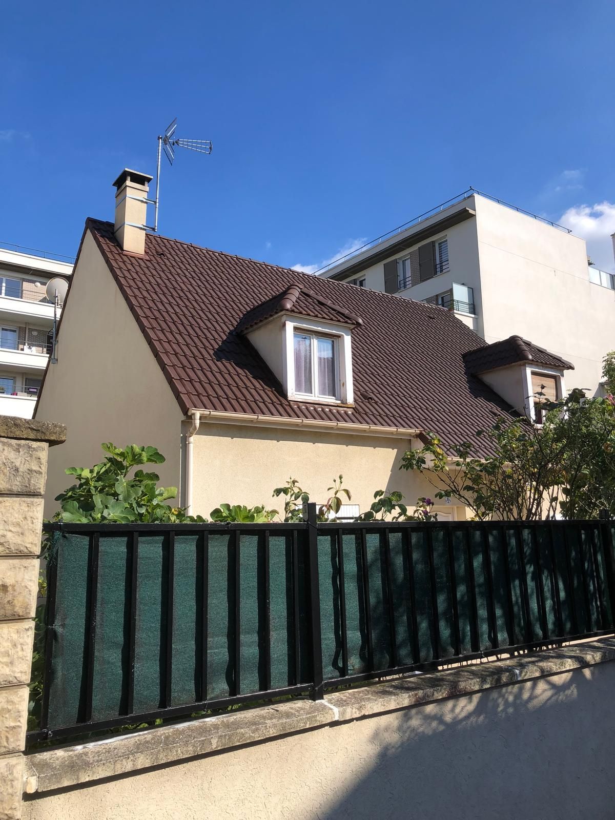 Une maison couleur crème avec un toit brun pentu, derrière une clôture métallique verte, sous un ciel bleu.