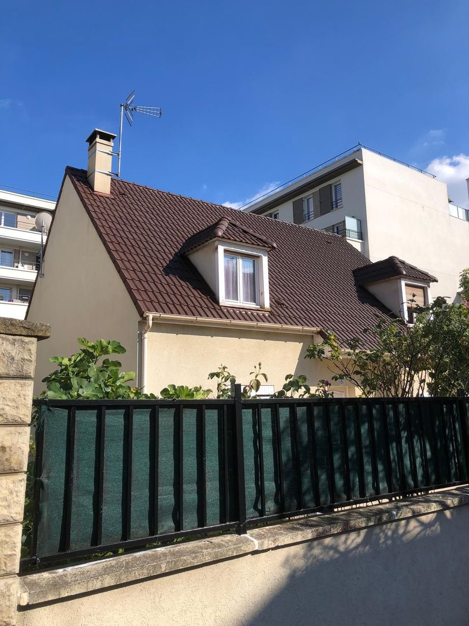 Une maison couleur crème avec un toit brun pentu, derrière une clôture métallique verte, sous un ciel bleu.