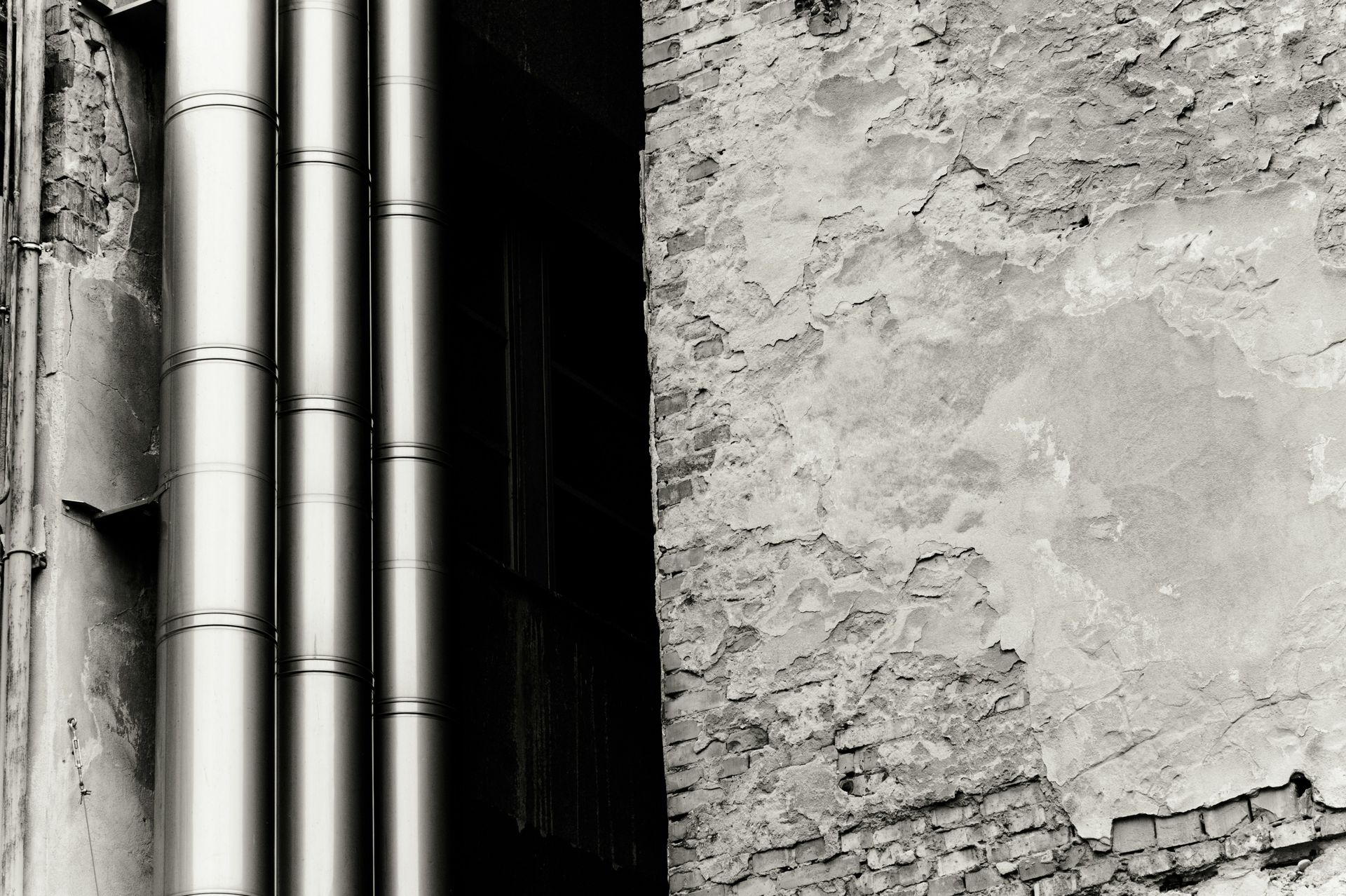 Des tuyaux métalliques verticaux bordent un mur à la texture rugueuse, en noir et blanc.