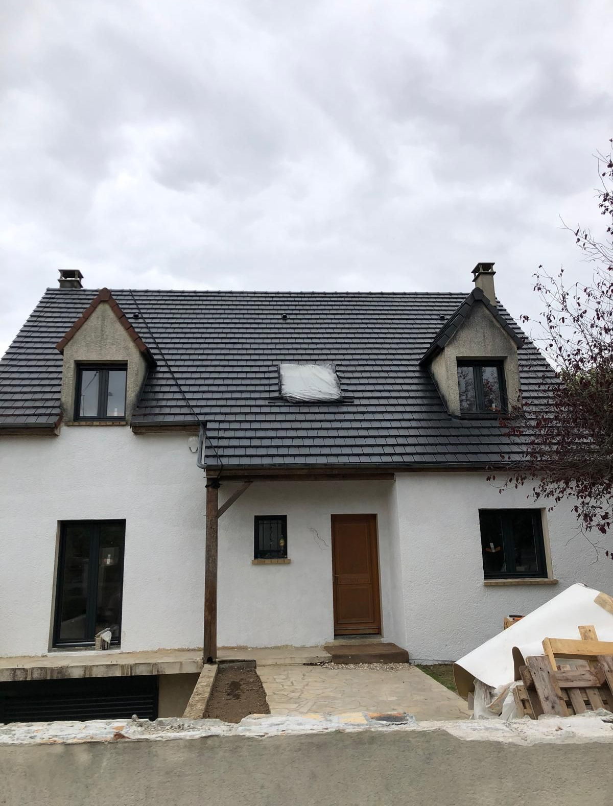 Maison blanche au toit gris et aux lucarnes sous un ciel nuageux, avec des matériaux de construction devant.