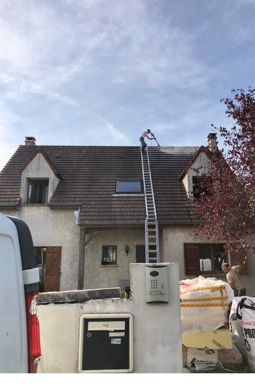 Extérieur d'une maison avec toit de tuiles, mur blanc, boîte aux lettres et lampadaire sous un ciel nuageux.