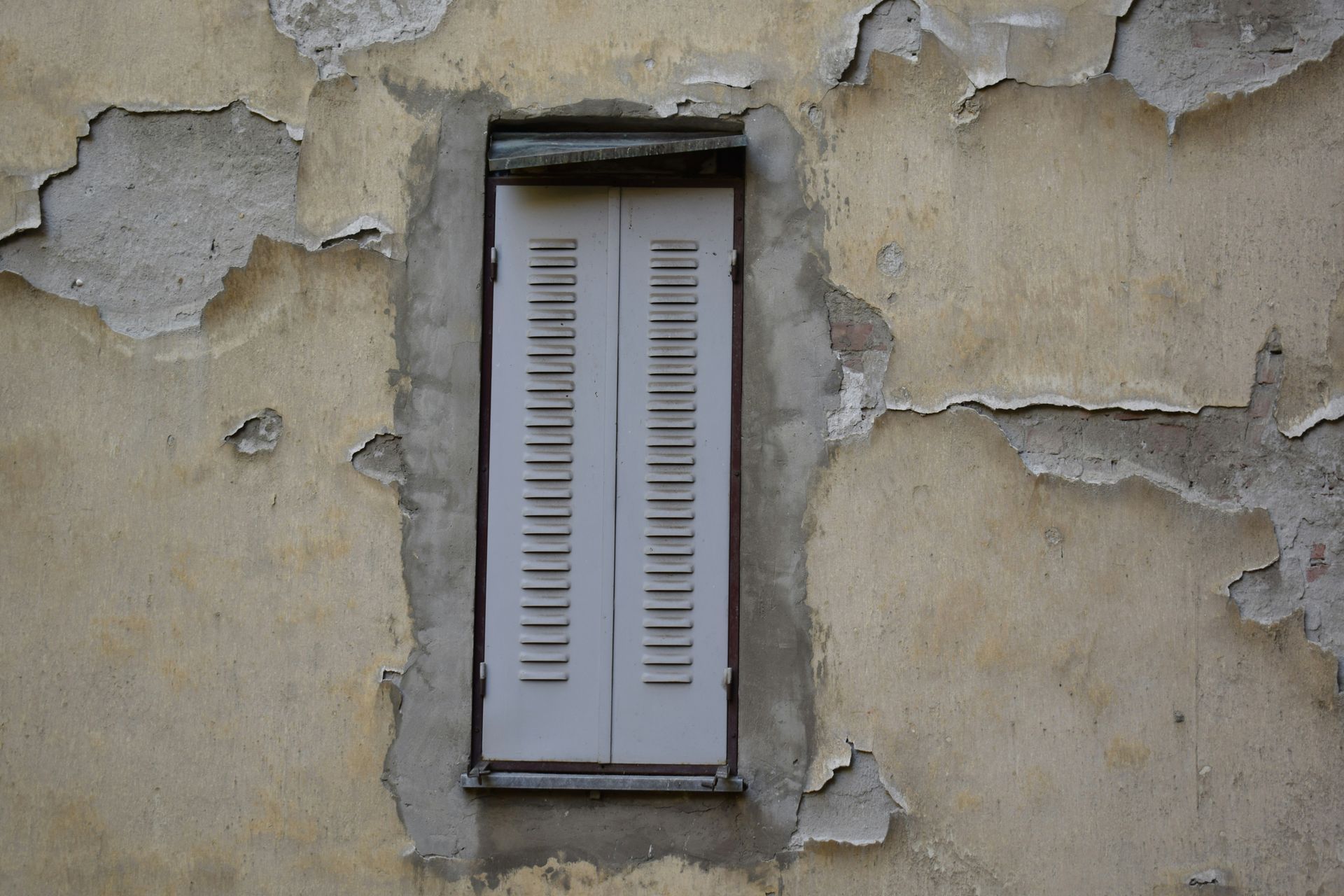 Fenêtre à volets blancs sur un mur beige délavé aux enduits écaillés.