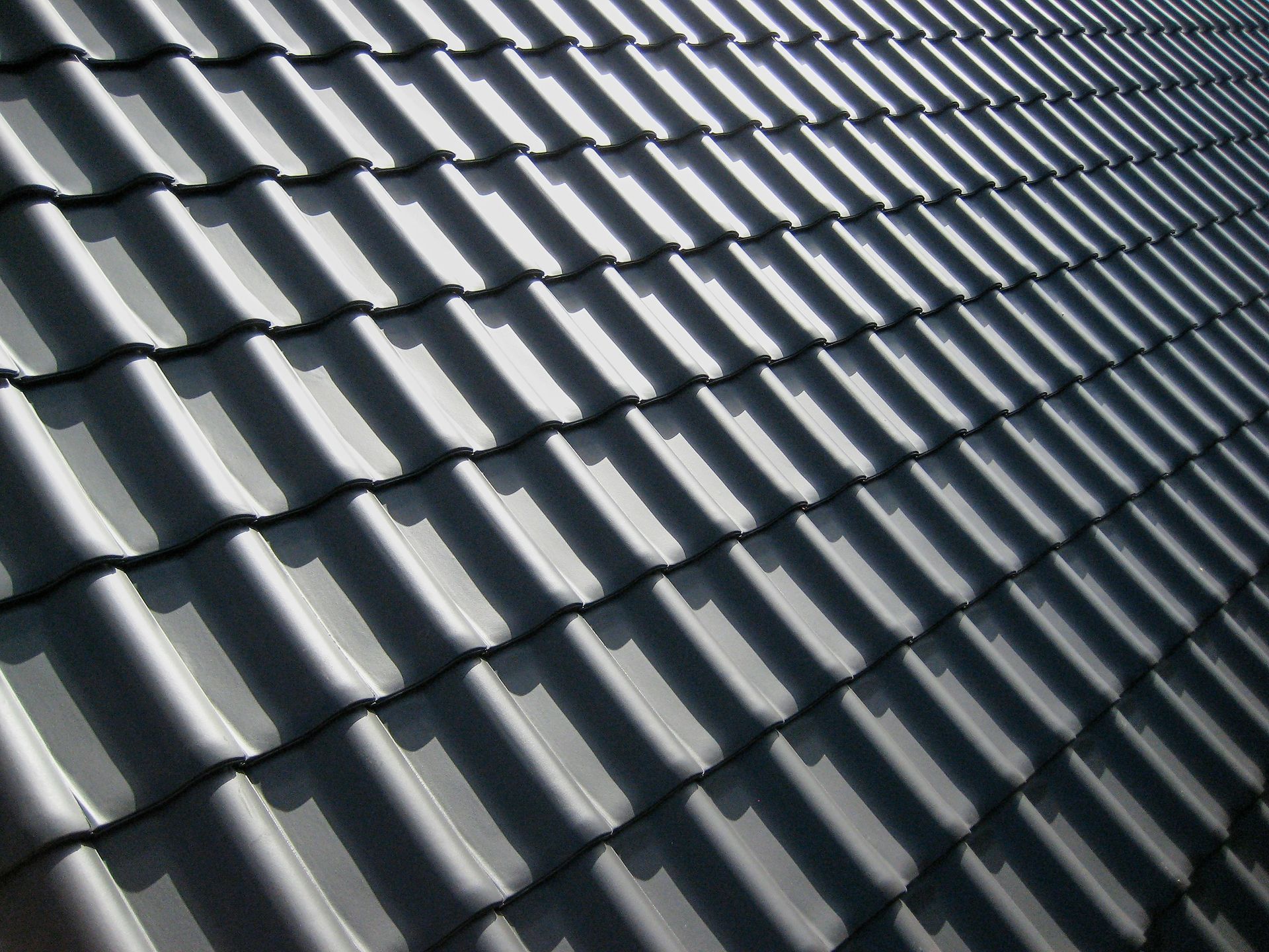 Gros plan sur des tuiles de toit en métal gris disposées en rangées diagonales, avec la lumière du soleil et les ombres.