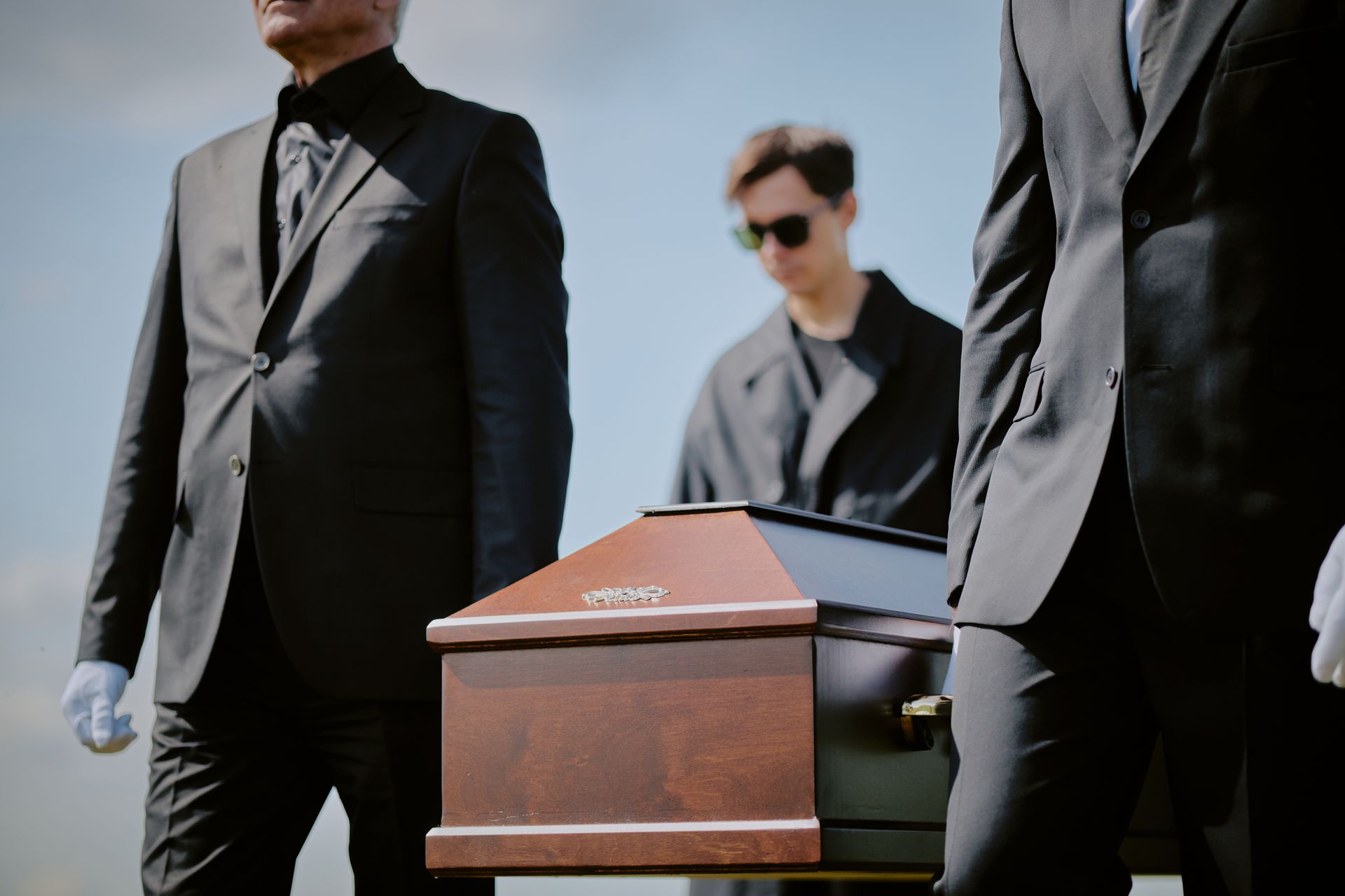 Des hommes en costumes noirs portent un cercueil en bois à l'extérieur, suivis d'une personne portant des lunettes de soleil.