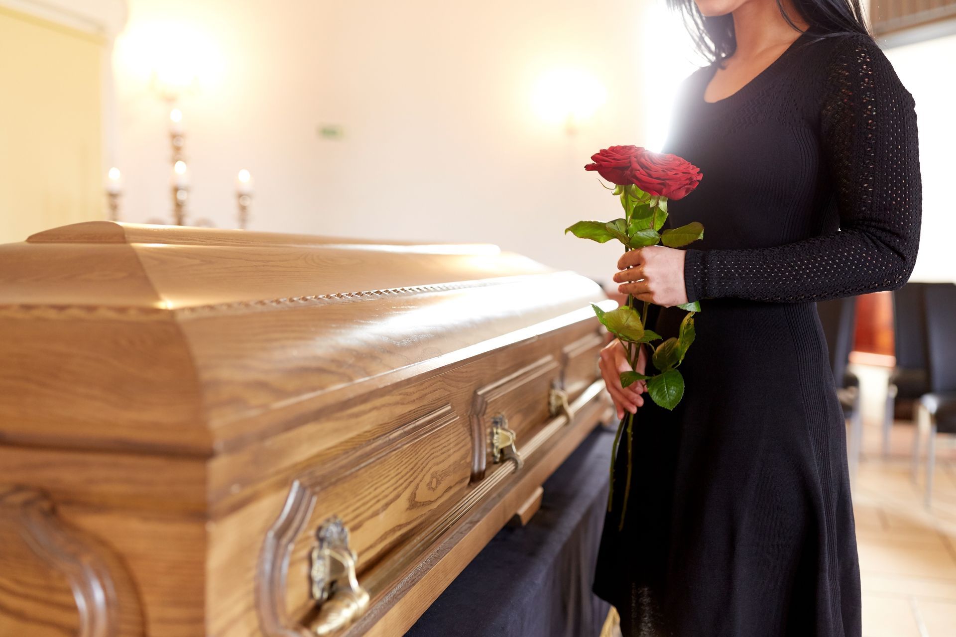 Femme en robe noire tenant une rose rouge, debout près d'un cercueil en bois fermé dans une pièce bien éclairée.