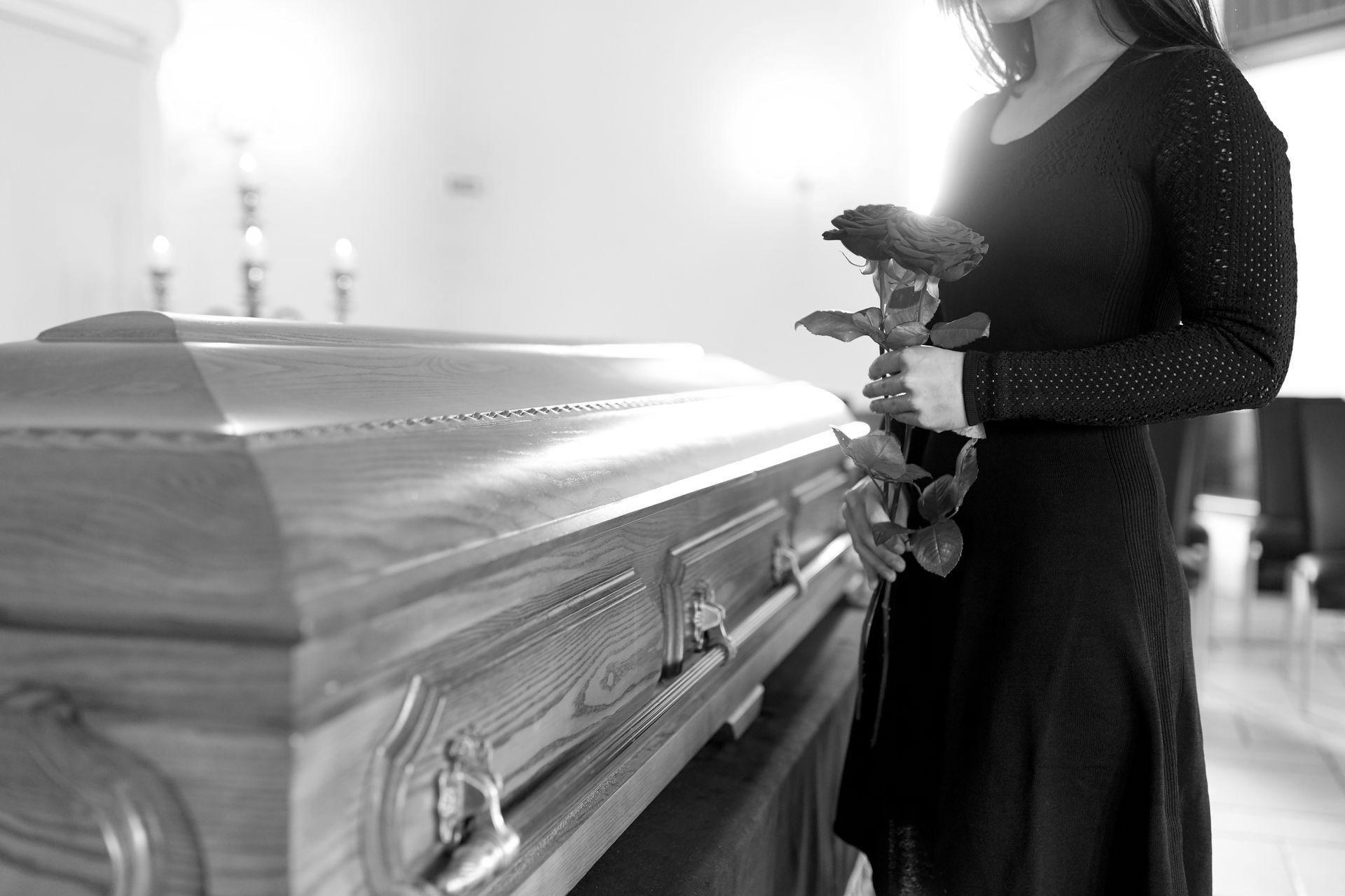 Femme en noir tenant une rose rouge, debout près d'un cercueil fermé dans une maison funéraire.