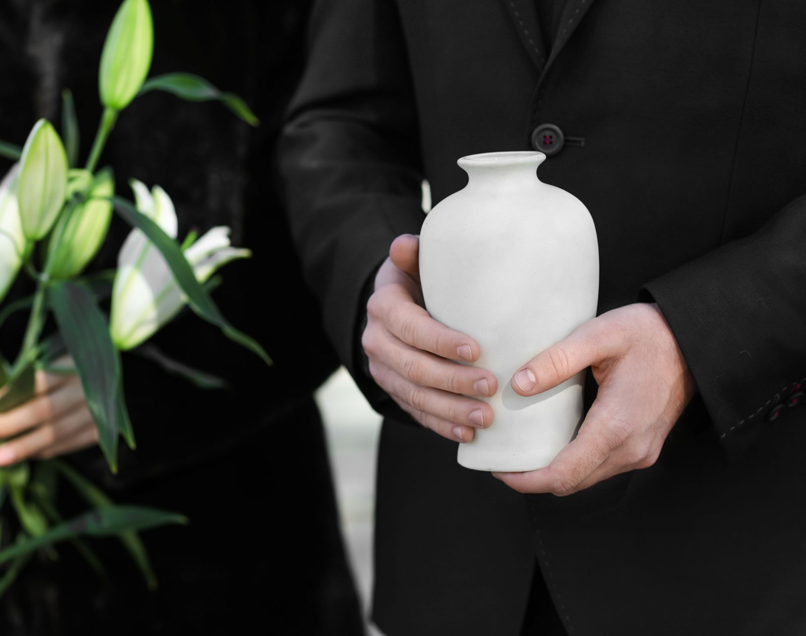 Un homme en costume noir tient une urne funéraire blanche ; une femme en noir tient des lys.