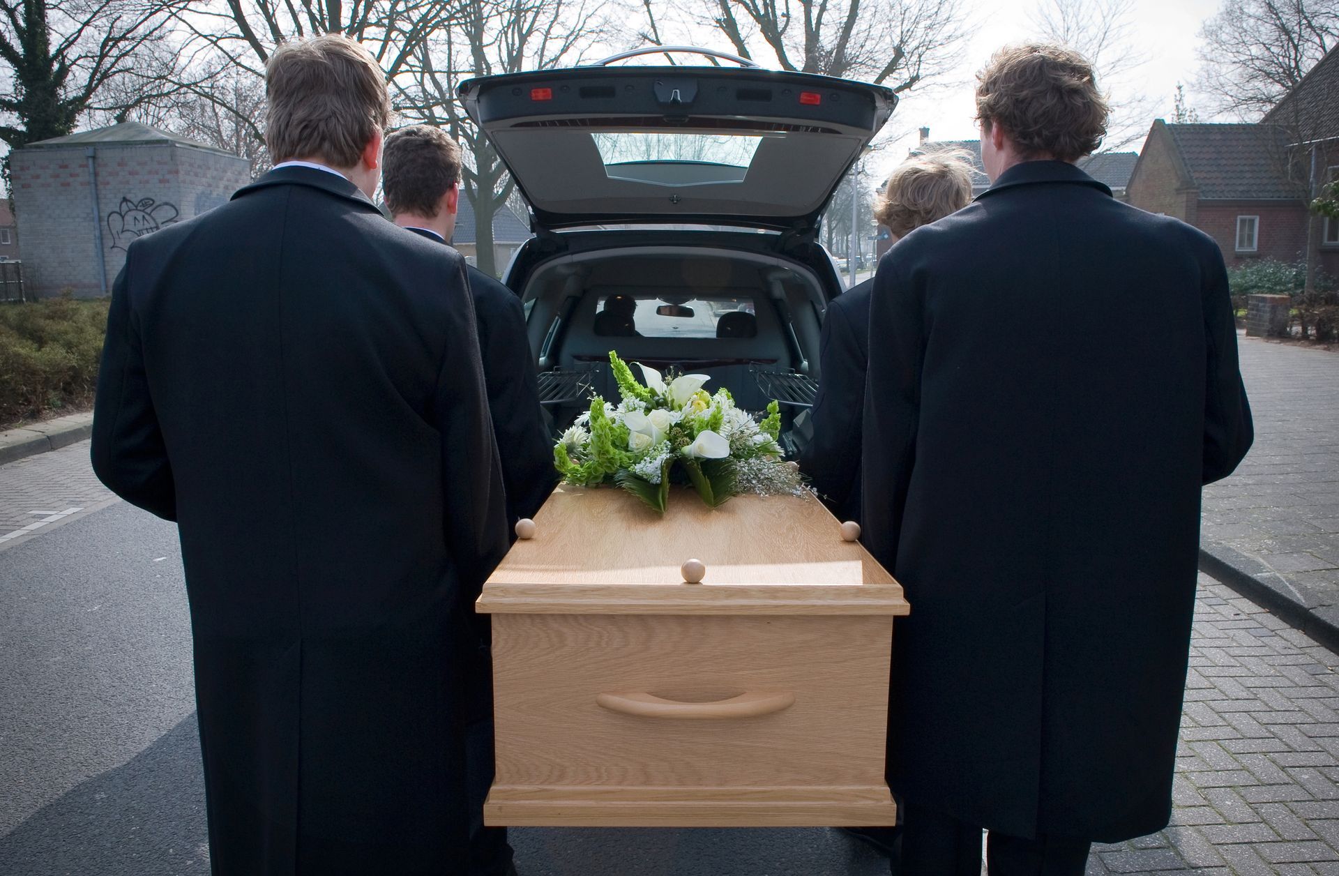 Des porteurs transportent un cercueil en bois vers une voiture ; des fleurs au sommet ; décor extérieur.