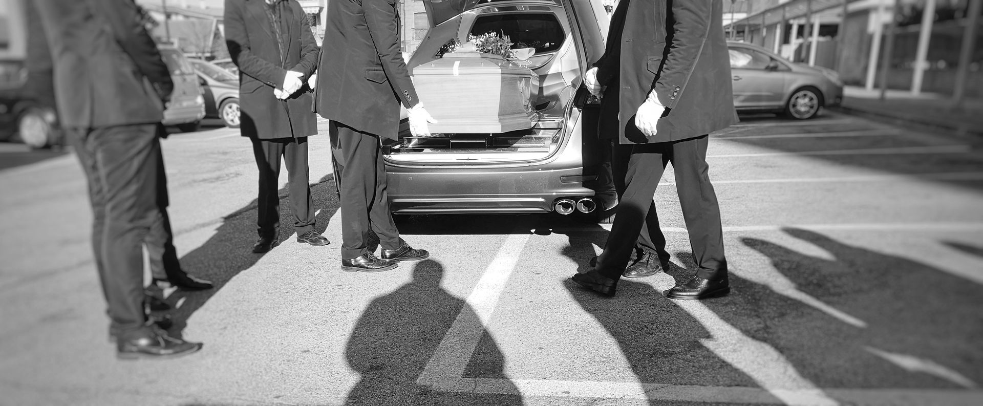 Des hommes en costume soulèvent un cercueil d'un corbillard sur un parking. La lumière vive du soleil projette de longues ombres.