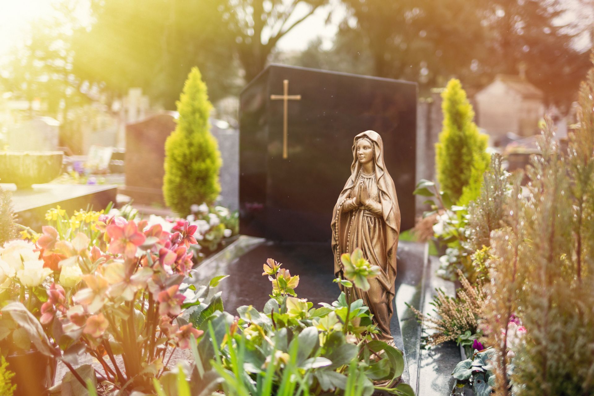 Scène de cimetière avec une pierre tombale noire, une statue en bronze et des fleurs colorées.