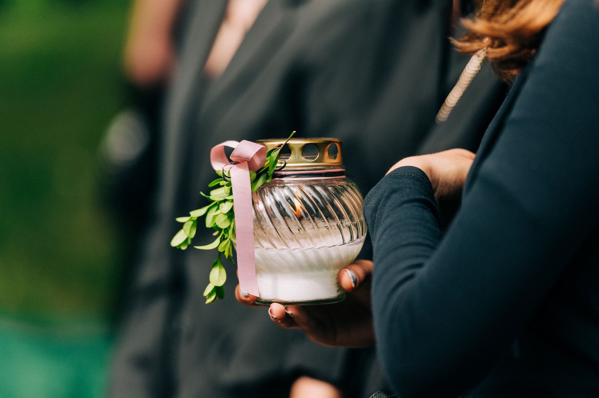 Personne tenant une bougie en verre avec un ruban rose et de la verdure ; arrière-plan flou.