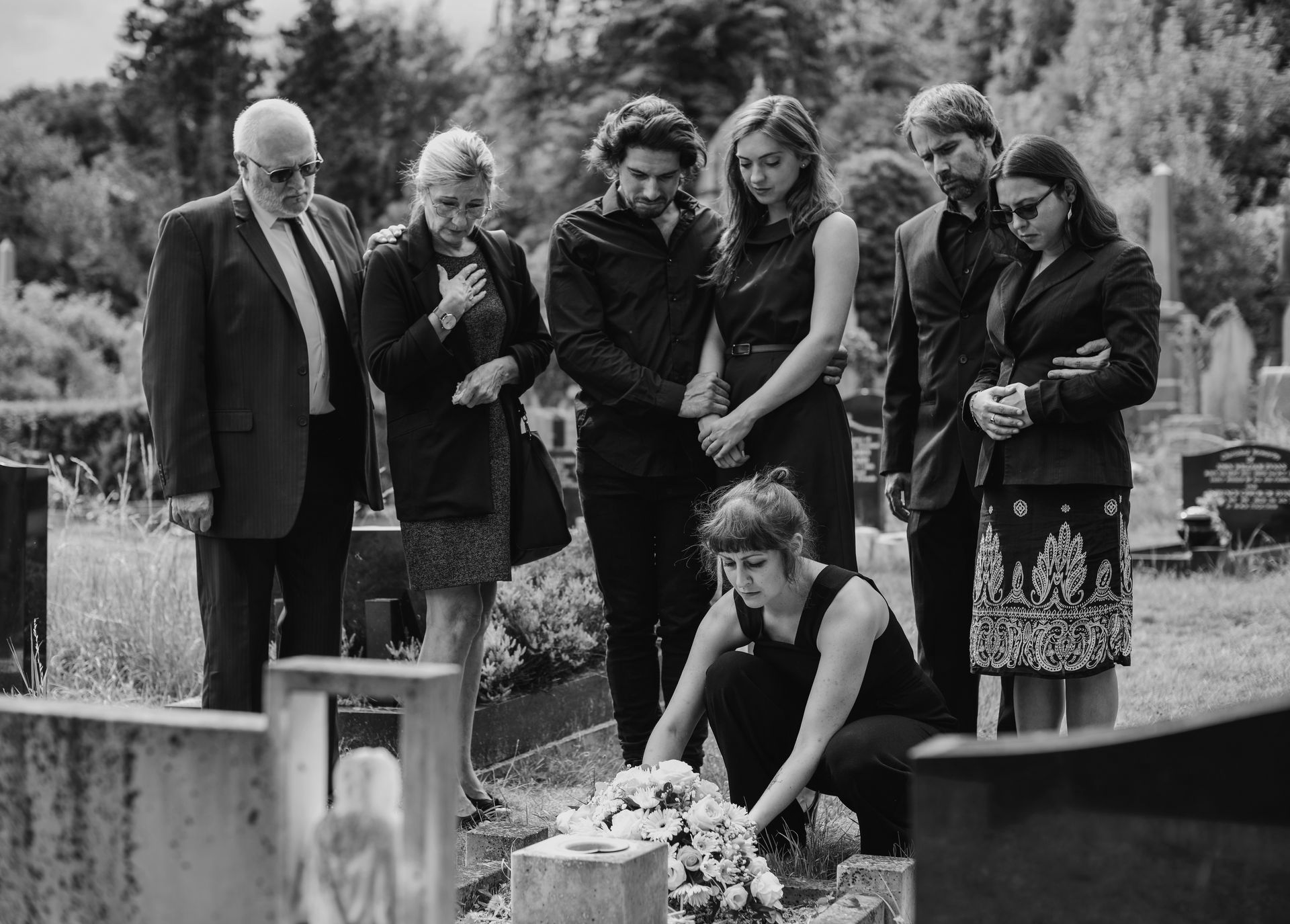 Des personnes en tenue noire sur une tombe ; une personne dépose des fleurs sur la tombe, d'autres pleurent.
