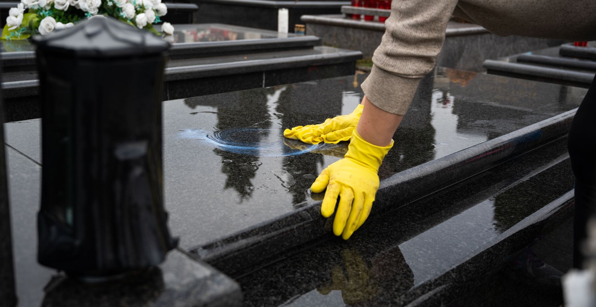 Une personne portant des gants jaunes nettoie une pierre tombale en granit noir dans un cimetière.