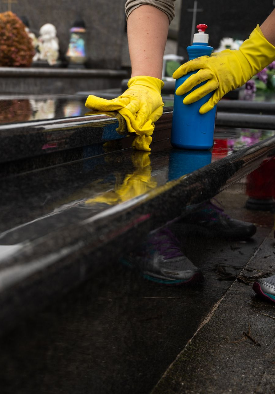 Une personne portant des gants jaunes nettoie une pierre tombale noire avec une bouteille bleue de nettoyant.