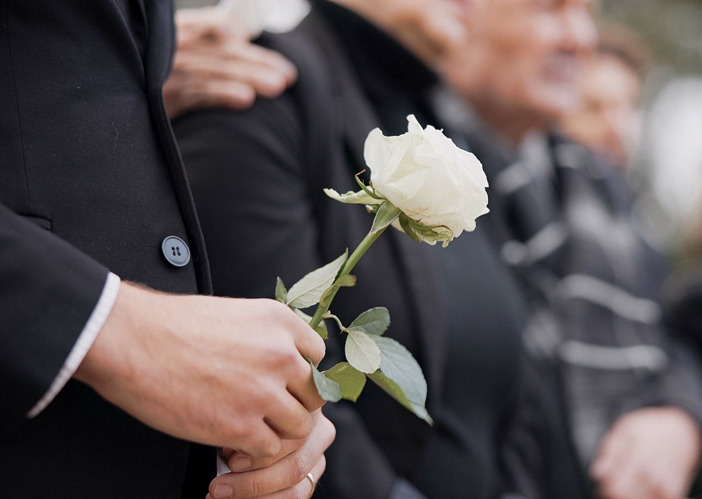 Un homme en costume noir tient une rose blanche, debout avec d'autres, lors d'un enterrement.
