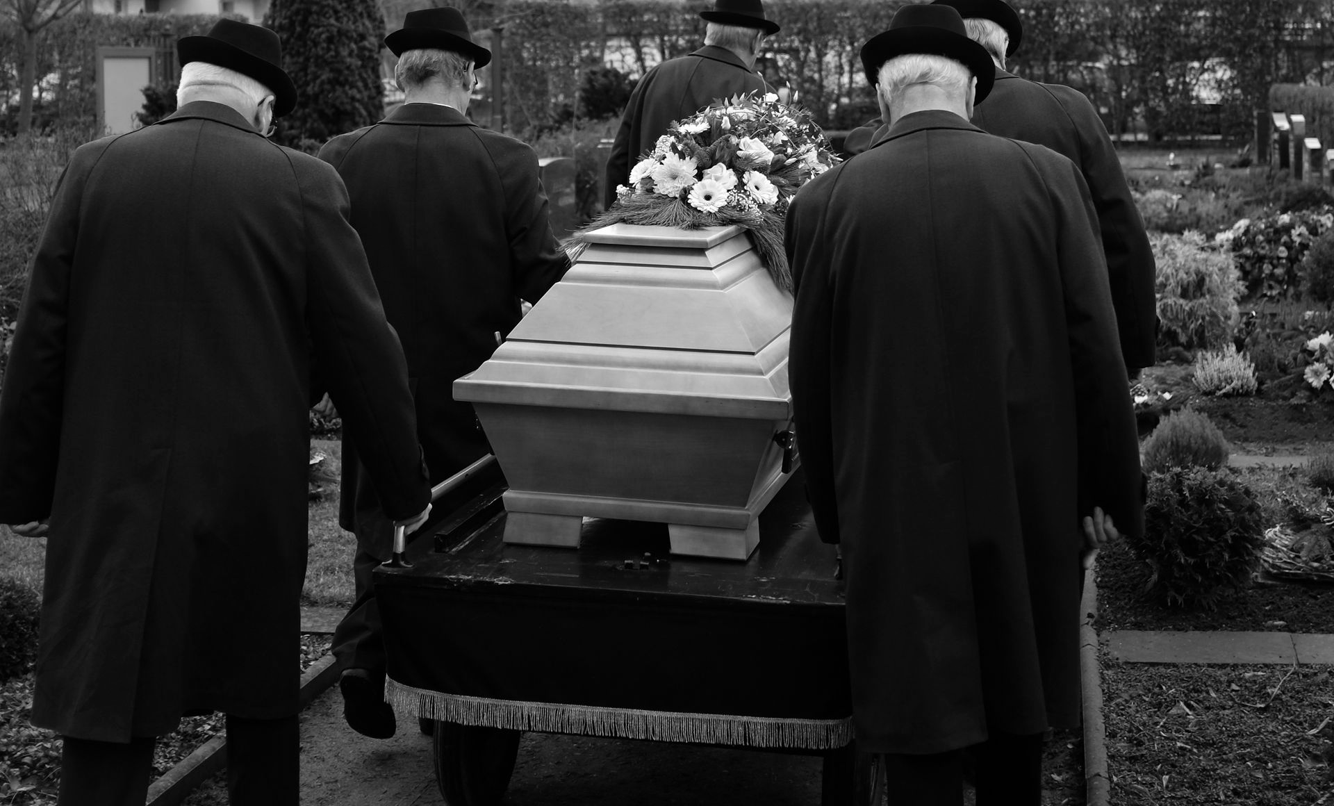 Des hommes en costumes noirs portent un cercueil avec des compositions florales dans un cimetière.
