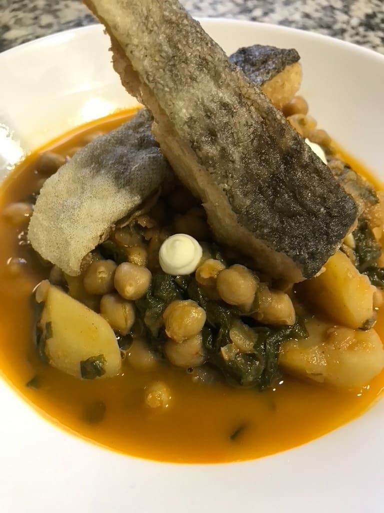 Bol de estofado de garbanzos cubierto con filete de pescado crujiente y verduras en caldo rojo.