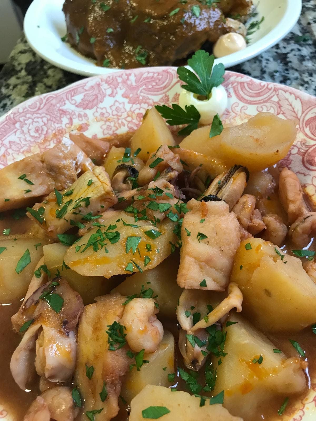 Guiso abundante con patatas, judías y hierbas en un plato estampado, con otro plato al fondo.