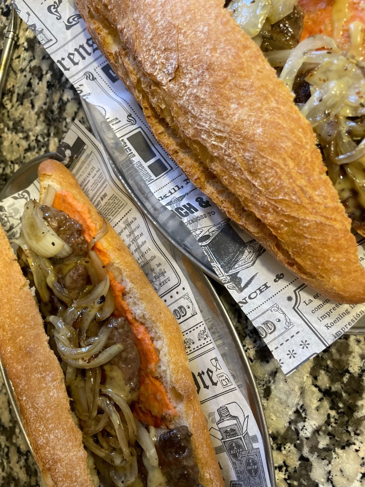 Dos sándwiches sobre papel de periódico en una encimera de granito, con el pan tostado y el relleno de carne a la vista.