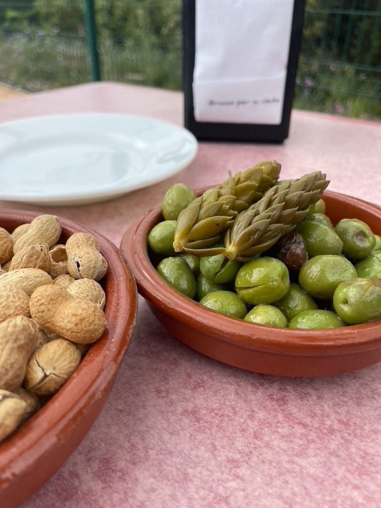 Cuencos de frutos secos tostados y aceitunas verdes con espárragos en una mesa al aire libre.