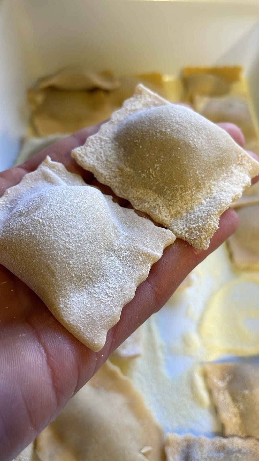 Una mano sostiene dos pasteles cuadrados espolvoreados con azúcar, con una bandeja de pasteles similares al fondo.