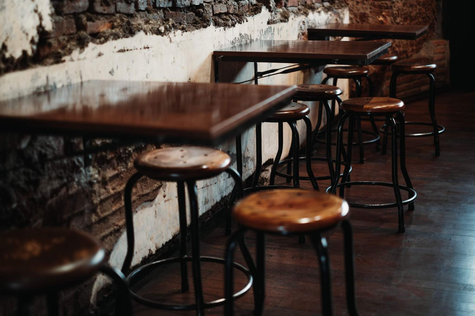 Cafetería vacía con mesas de madera, taburetes redondos y una pared de ladrillo desgastada.