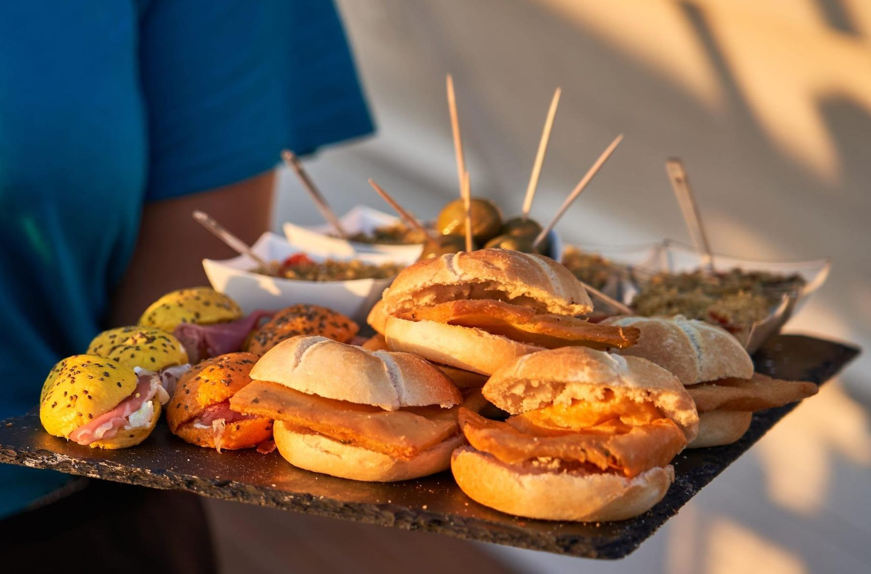 Plato con sándwiches variados y brochetas sobre una mesa al aire libre al atardecer.