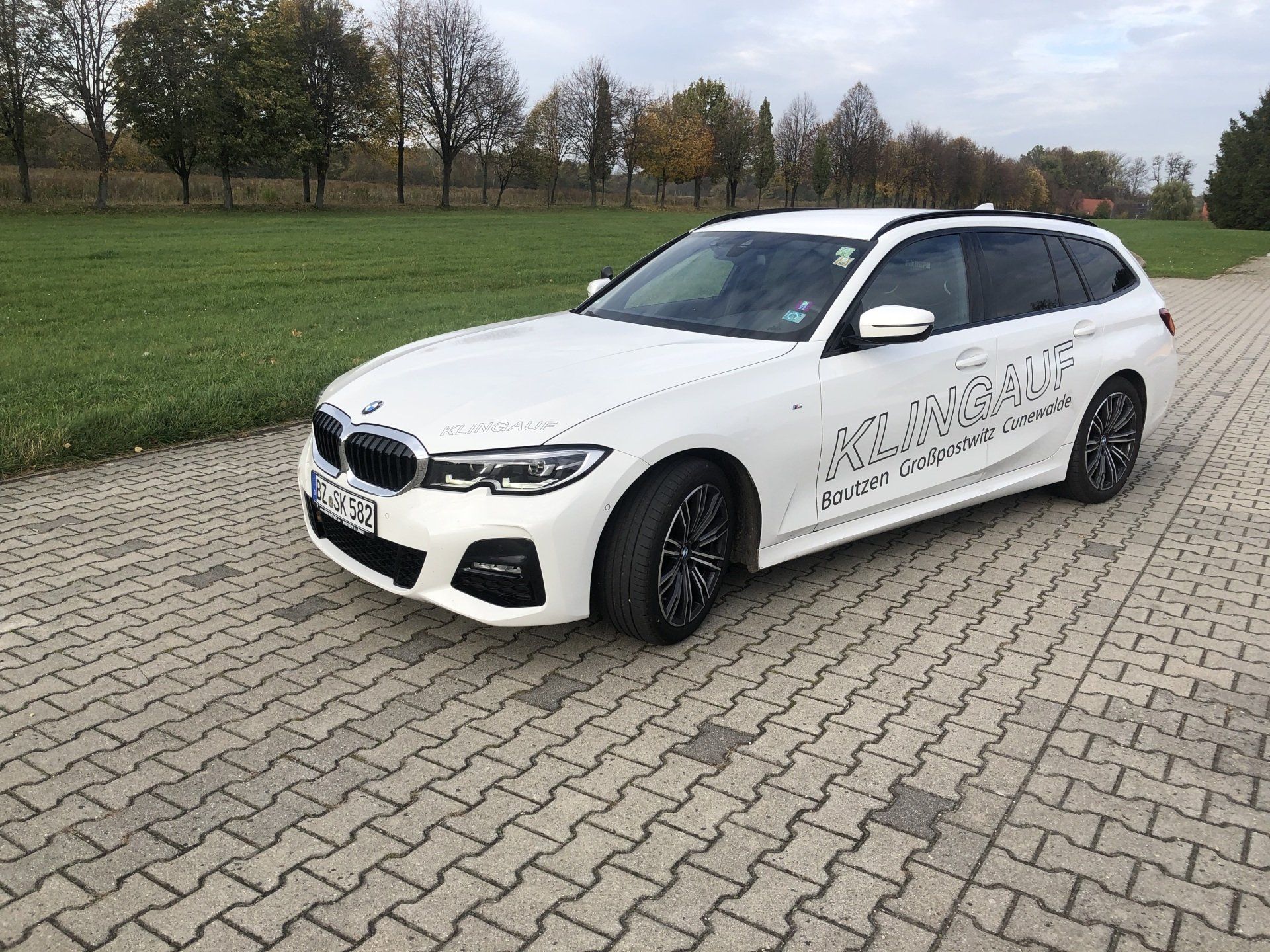 Weißer BMW-Kombi, geparkt auf einer Ziegelstraße mit grünem Gras und Bäumen im Hintergrund.
