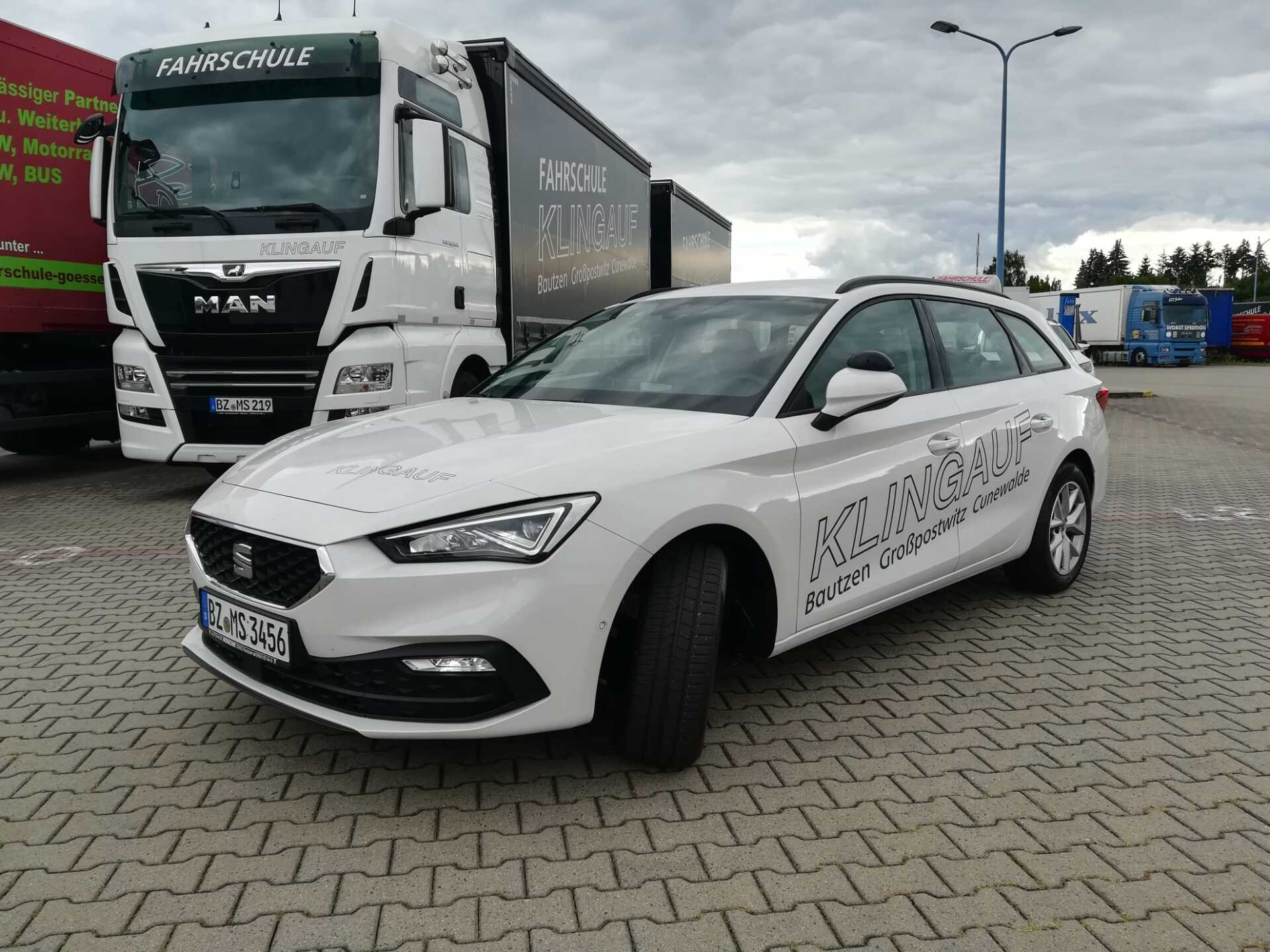 Weißes Auto parkt neben einem LKW auf einem Parkplatz. Grauer Bürgersteig, bewölkter Himmel.