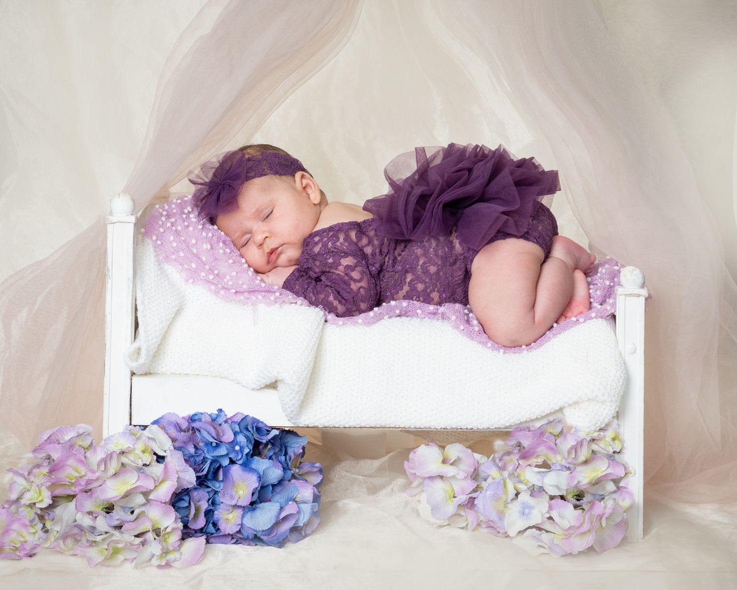 Bebé durmiendo con un vestido morado en una cama blanca con flores de lavanda a su alrededor.