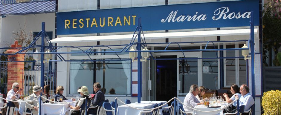La gente cena en mesas en el patio al aire libre del restaurante azul y blanco María Rosa en un día soleado.