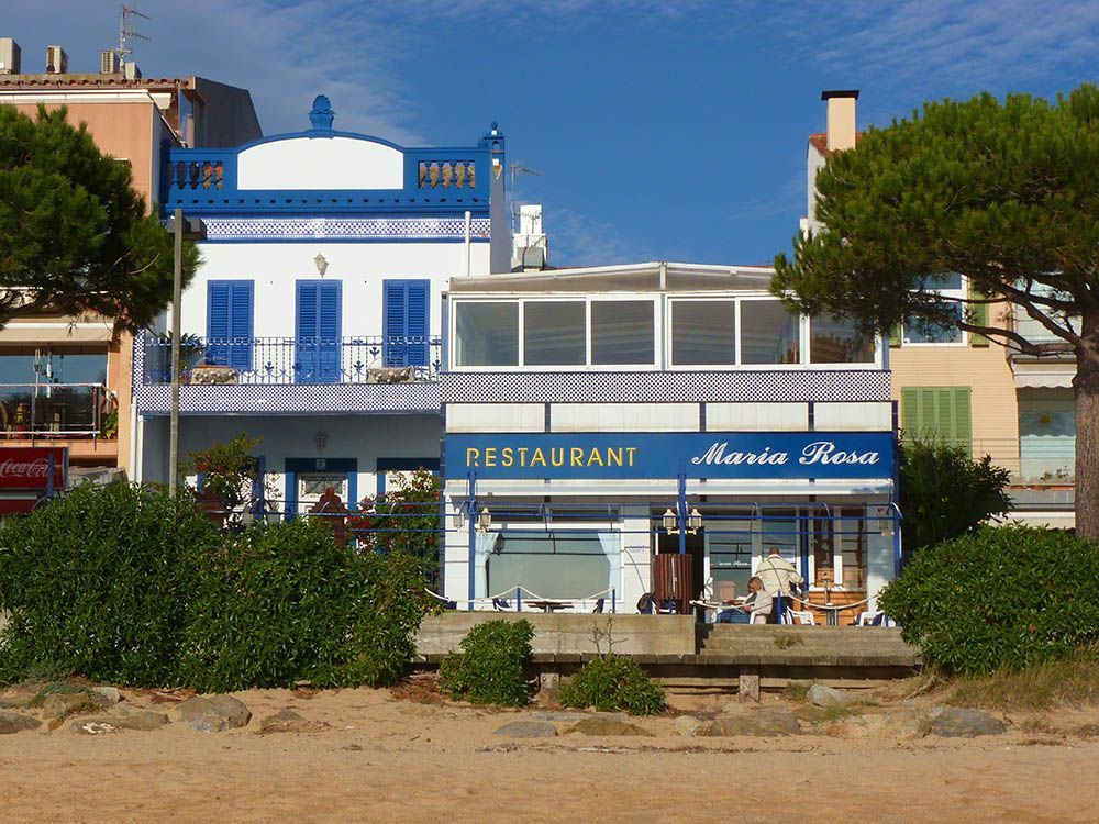 Una vista soleada de la playa de un edificio de estilo colonial con detalles en azul y blanco, al lado de un restaurante moderno llamado María Rosa.